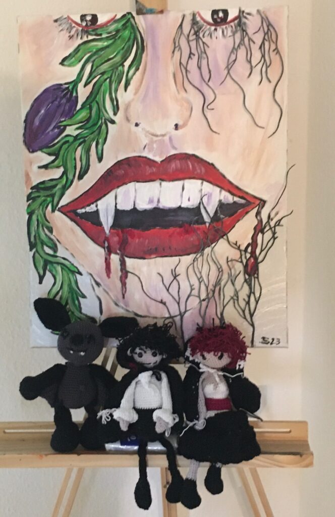Bild eines gemalten Vampirkopfes mit gehäkelten Vampiren und Fledermaus davor auf einer Staffelei